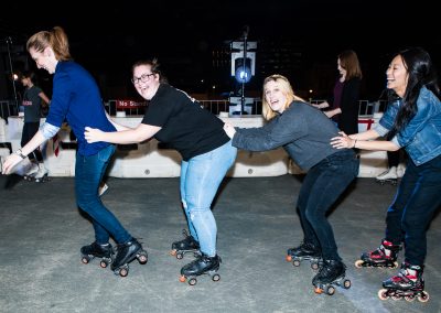 4 Women in a line holding onto each others back skating with Malt Shop Rollers at a Community Event.